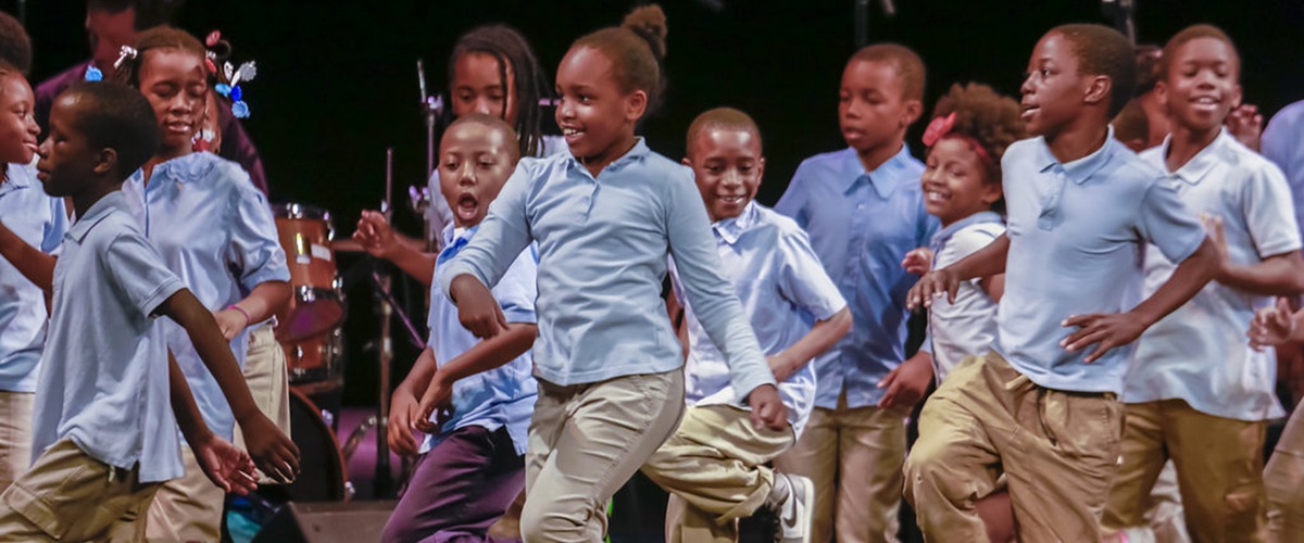 A group of school-aged children in school uniforms of light blue shirts and khaki  pants dance on stage