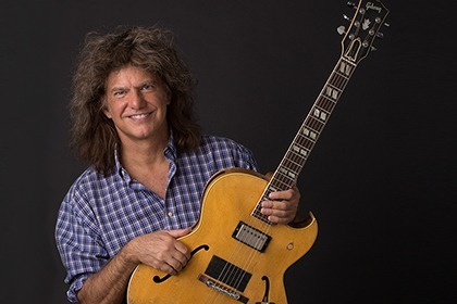 Pat Metheny Sits with an electric guitar on his lap smiling at the camera against a dark background