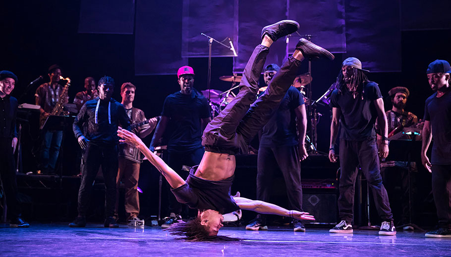 dancer posed on stage on his head while band watches in the background