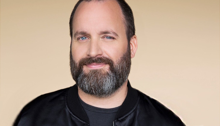 Tom Segura looking at camera in a black jacket with a beige background