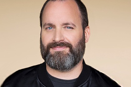 Tom Segura looking at camera in a black jacket with a beige background