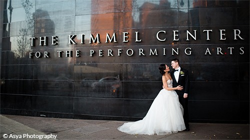 Monica and Andrew in wedding attire stand in front of the Kimmel Center Building Exterior